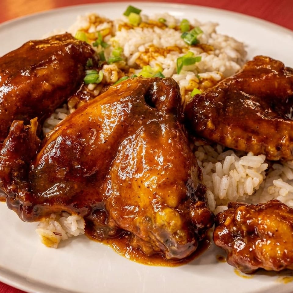 Glazed braised chicken pieces served over white rice with green onions on a white plate.